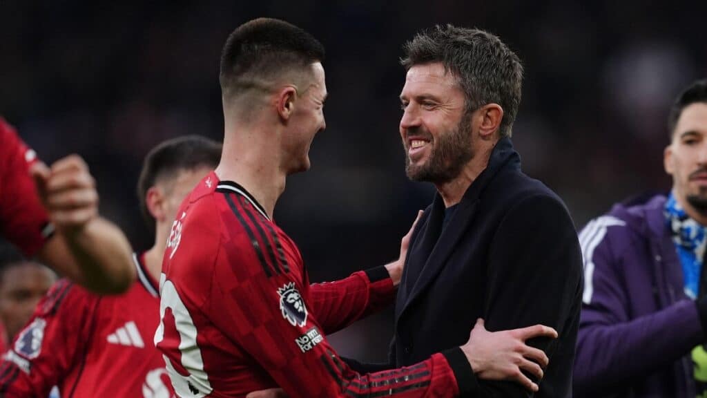 O técnico do Manchester United Football Club, Michael Carrick, parabeniza um de seus jogadores voltado para a esquerda, sorrindo e vestindo uma blusa preta. Carrick tem cabelo escuro, bigode e barba. Seu jogador está vestindo uma camisa vermelha, preta e branca do Man United, com outros jogadores visíveis em um campo de futebol