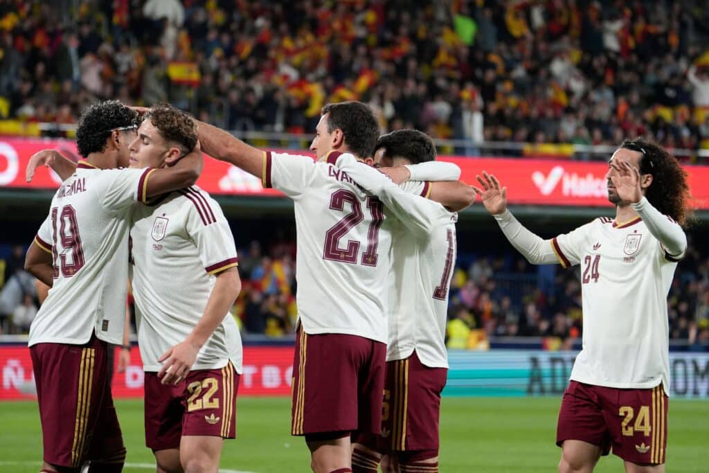 Mikel Oyarzabal comemora após marcar pela seleção espanhola, abraçado pelos companheiros de costas para o gol, com camisas brancas e shorts vermelhos, com os números do elenco à mostra