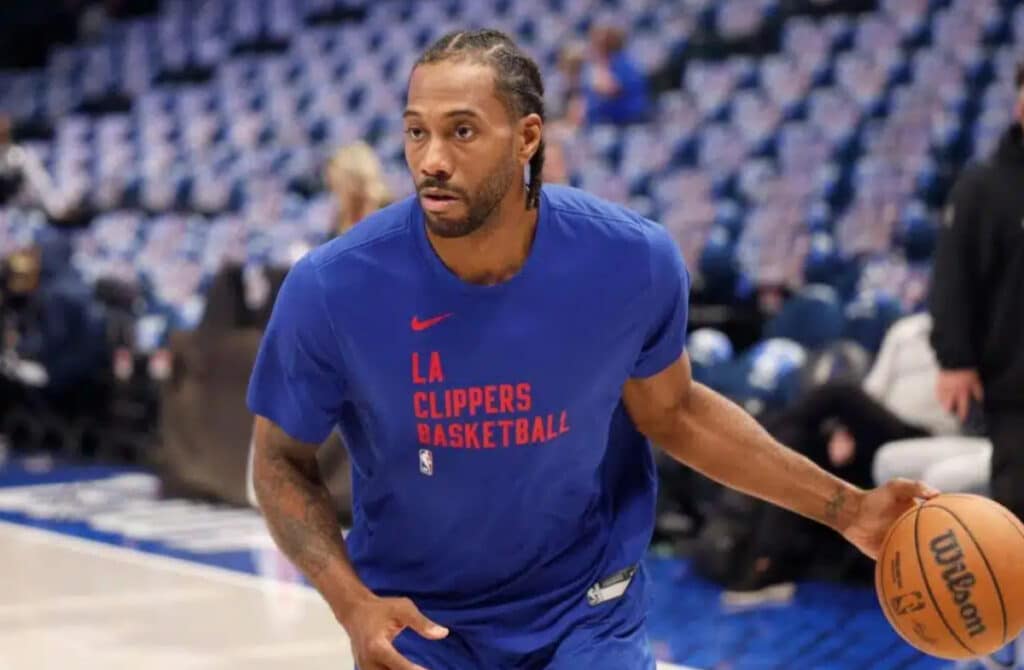 O jogador de basquete da NBA Kawhi Leonard segura uma bola laranja com a palavra Wilson em preto em uma quadra em frente a assentos vazios. Leonard está olhando para a direita e vestindo uma camiseta azul com as palavras Los Angeles Clippers em vermelho. As pessoas estão desfocadas, mas visíveis no fundo